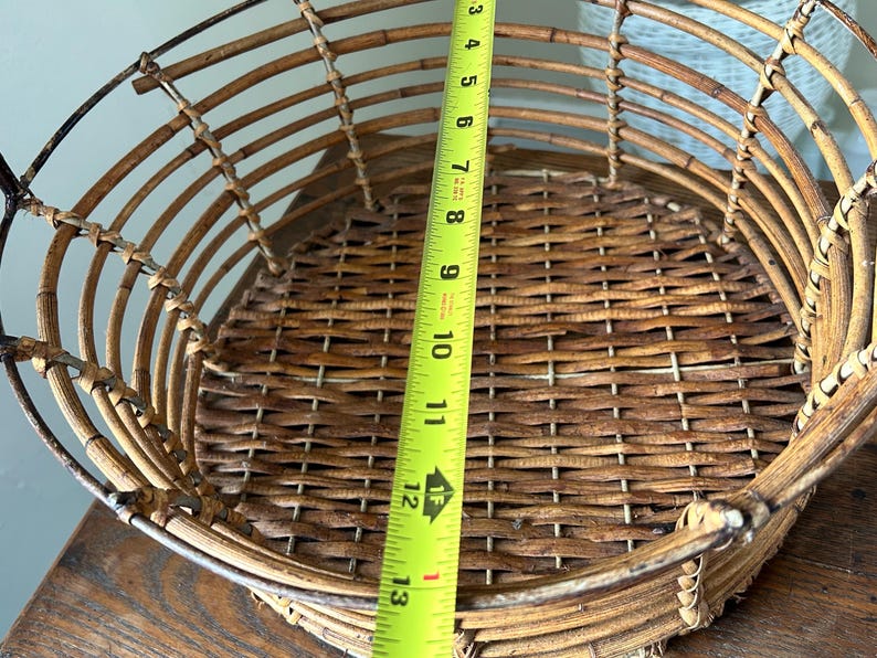 May include: A woven wicker basket with a metal frame. The basket is brown and has a circular shape. A measuring tape is placed next to the basket, showing a measurement of 13 inches.
