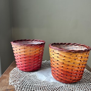 May include: Two woven baskets with a gradient of colors from red to orange and yellow. The baskets are lined with a clear plastic liner. They sit on a white, crocheted doily on a wooden table. The baskets are suitable for plants.