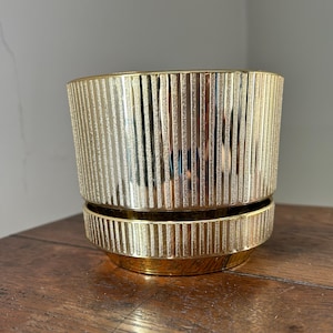 May include: A gold-coloured planter with a ribbed design. The planter has a cylindrical shape with a matching saucer. The metallic finish gives it a modern and elegant look. Ideal for displaying plants or as a decorative accent.