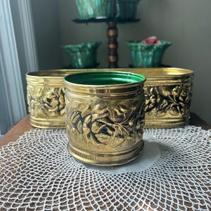 May include: A round brass planter featuring a raised floral relief pattern with a green interior, positioned in front of two larger oval brass planters that also display a matching embossed floral design and green interior finish.