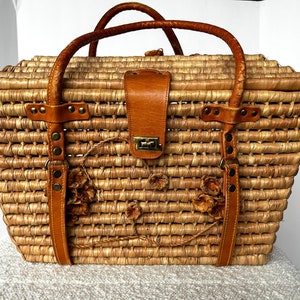 May include: A large woven straw tote bag with brown leather handles and straps. The bag has a decorative floral design and a brass clasp.