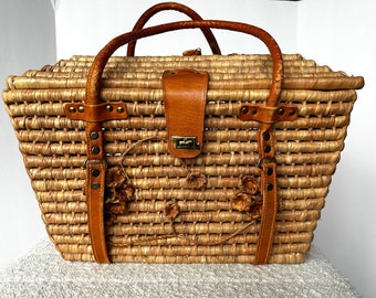 May include: A large woven straw tote bag with brown leather handles and straps. The bag has a decorative floral design and a brass clasp.