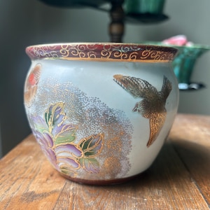 May include: A small, round porcelain planter featuring a white base with a dark red rim. The exterior is decorated with a gold-toned bird in flight, purple and green floral motifs, and a stippled gold texture pattern around the center.