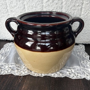 May include: A brown and beige glazed ceramic crock with two handles. The crock has a rounded body and a wide opening.