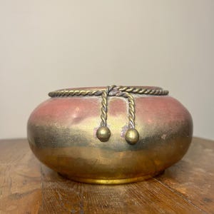May include: A decorative, antique-style bowl with a rounded shape. The bowl has a metallic finish, with a gradient of colors from gold to reddish-brown. It features a rope-like trim around the rim and two dangling gold spheres.