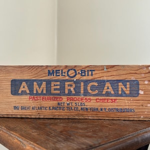May include: A vintage wooden cheese box with the text "MEL-O-BIT AMERICAN PASTEURIZED PROCESS CHEESE" in blue and red lettering. The box is made of weathered wood and has the text "NET WT. 5 LBS." and "THE GREAT ATLANTIC & PACIFIC TEA CO, NEW YORK, N.Y., DISTRIBUTORS".