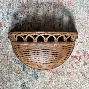May include: A brown, semi-circular woven basket with a decorative arched top. The basket is made of natural-colored wicker and is designed to be mounted on a wall. The basket's woven pattern is visible, and it appears to be empty.