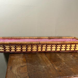 May include: Rectangular wicker basket with a red and white gingham fabric lining. The basket is made of woven light brown and dark brown wicker. The basket is sitting on a wooden surface.