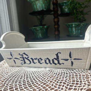May include: Cream ceramic bread basket, rectangular with handles. The word "Bread" is written in dark blue gothic lettering. The basket sits on a lace doily. The basket is likely for kitchen use.
