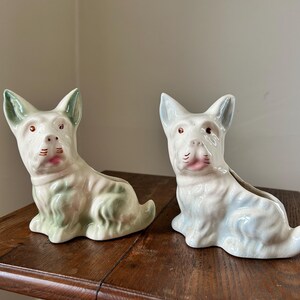 May include: Two ceramic dog figurines, each in a seated position. The dogs are white with green and blue accents, pink mouths, and brown eyes. They are sitting on a wooden surface.