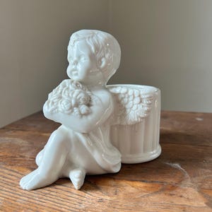 May include: A white ceramic cherub planter. The cherub is seated, holding a bouquet of flowers, with a small planter behind it. The planter has a fluted design and angel wings. The cherub is on a wooden surface.