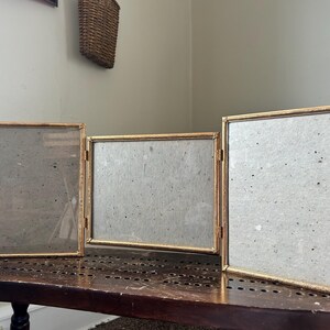 May include: A gold-framed, three-panel picture frame set. The frames are hinged together, allowing them to stand upright. The frames have a simple, elegant design, with a gold finish. The frames are empty, ready to display photos or artwork.