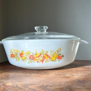 May include: White casserole dish with a clear glass lid. The dish features a colorful vegetable design around the base, including tomatoes, squash, and carrots. The dish is sitting on a wooden surface.