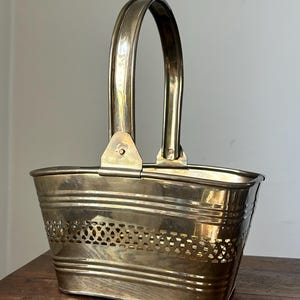 May include: A vintage brass basket with a curved handle. The basket has a rectangular shape with horizontal lines and a decorative pierced pattern. The handle is attached with small brass brackets. The basket is sitting on a wooden surface.
