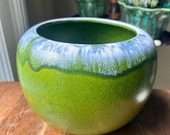 May include: A round, ceramic planter with a glossy finish. The planter features a gradient of colors, transitioning from a vibrant green base to a blue and white rim. The interior is a deep green. Other planters are visible in the background.