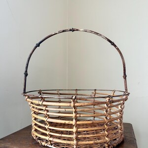 May include: A woven wicker basket with a brown metal handle. The basket is round and has a natural, rustic look.