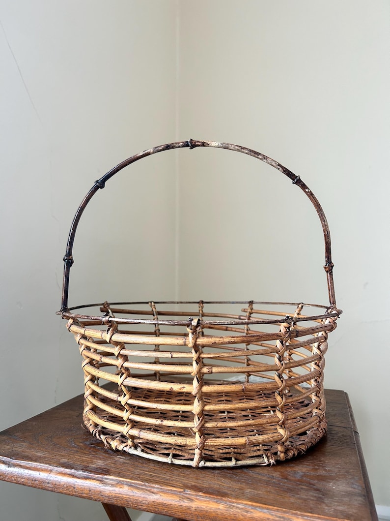 May include: A woven wicker basket with a brown metal handle. The basket is round and has a natural, rustic look.