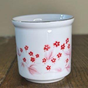 May include: White ceramic cup with a gold rim, adorned with a floral pattern of red flowers and pink leaves. The cup has a cylindrical shape and sits on a wooden surface. A decorative piece for the kitchen or dining room.