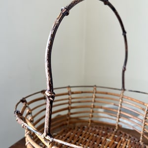 May include: A woven wicker basket with a brown, metal handle. The basket is oval shaped and has a woven pattern.
