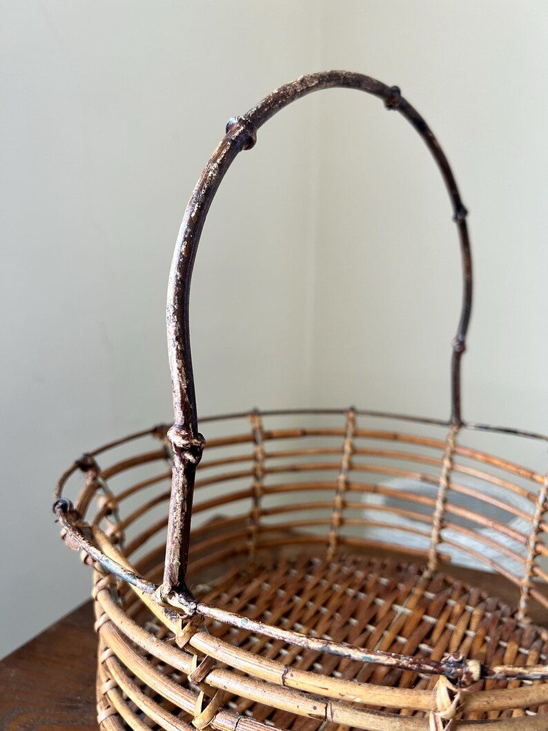 May include: A woven wicker basket with a brown, metal handle. The basket is oval shaped and has a woven pattern.
