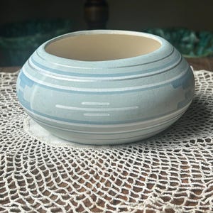 May include: A light blue ceramic bowl with horizontal stripes in white and light blue. The bowl has a round shape and a beige interior. It sits on a white, crocheted doily, suggesting a decorative or home decor item.