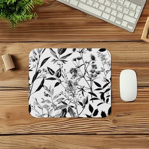 May include: A rectangular mousepad with a white background and a black and white botanical design featuring various leaves and floral elements. The mousepad is surrounded by a wooden desk, a computer mouse, a keyboard, and a small plant.