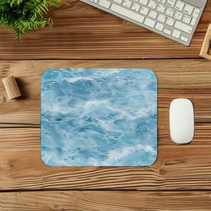 May include: A rectangular mousepad featuring a blue and white watercolor design of ocean waves. The mousepad is on a wooden desk with a white computer mouse, keyboard, and a small green plant.