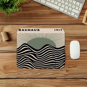 May include: A rectangular mousepad featuring a Bauhaus design. The design includes a green sun, radiating lines, and black and white wavy lines. The word "BAUHAUS" and the number "1917" are printed at the top.