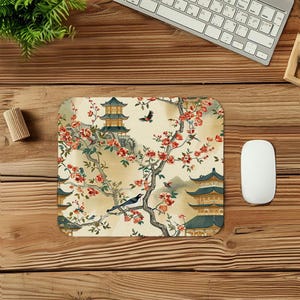 May include: A square mouse mat with a Japanese-inspired design. The design features pagodas, flowering branches with red blossoms, and birds against a cream background. The mat is surrounded by a wooden desk, a keyboard, and a computer mouse.