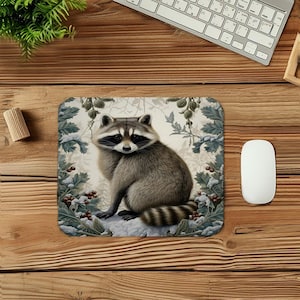 May include: A rectangular mouse mat with a detailed illustration of a raccoon. The raccoon has grey and brown fur, a black mask, and a striped tail, set against a backdrop of winter foliage and snow.