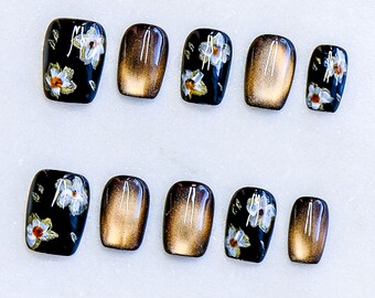Könnte beinhalten: Ein Set aus zehn künstlichen Nägeln. Einige Nägel sind schwarz mit weißen und orangefarbenen Blumenmustern, während andere einen Farbverlauf von Braun und Gold aufweisen. Der Text "DeCOLORS.Nails Art" befindet sich unten.