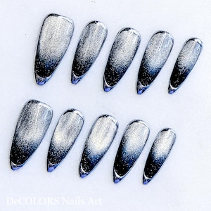 Puede incluir: Un conjunto de diez uñas postizas plateadas y azul marino. Las uñas tienen un acabado brillante con un efecto de purpurina que se desvanece de plata a azul marino. Las puntas de las uñas son puntiagudas y tienen un borde morado. El texto "DeCOLORS Nails Art" está en la parte inferior.
