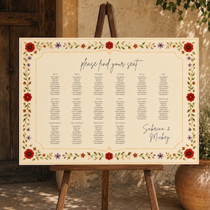 May include: A rectangular wedding seating chart on a wooden easel with a cream background and a floral border of red poppies and small purple flowers. The text reads please find your seat, lists tables one through sixteen, and Sabrina & Mickey.