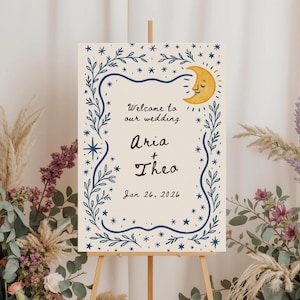 May include: A cream-colored wedding welcome sign on a wooden easel. The sign features a blue floral border, stars, and a smiling yellow moon. Text reads "Welcome to our wedding Aria + Theo Jan 26, 2026."