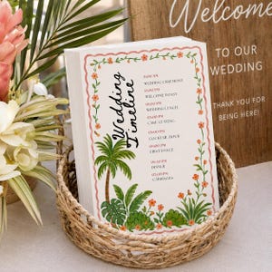 May include: A stack of wedding timeline cards in a woven basket. The cards feature a tropical design with a palm tree and floral border. The text on the cards includes the wedding timeline and the words "Wedding Timeline". A wooden sign that says "Welcome to our wedding" is in the background.