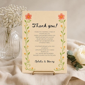 May include: A rectangular thank you card on cream paper featuring a floral border with orange flowers and green vines. The text reads: Thank you! Thank you for being a part of our special day. Ophelia & Henry. The card is held by a gold stand.