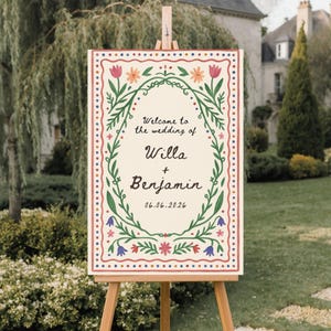 Può includere: Un cartello di benvenuto per matrimoni su un cavalletto di legno. Il cartello presenta uno sfondo color crema con un bordo floreale e il testo "Welcome to the wedding of Willa + Benjamin 06.06.2026."