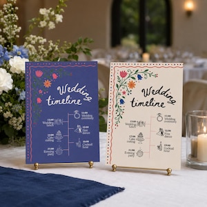 May include: Two wedding timeline signs on gold stands. One is navy blue with pink and orange floral accents, the other is cream with colorful floral accents. Both signs have the words "Wedding timeline" and a schedule.