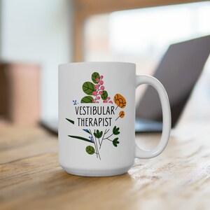 May include: White ceramic mug with the text "VESTIBULAR THERAPIST" in black, accompanied by a floral design in green, pink, and orange. The mug has a large handle and is likely used for hot beverages.