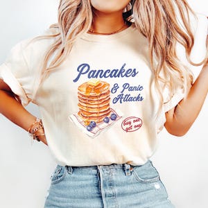 May include: A light beige t-shirt with a graphic of a stack of pancakes with blueberries and a butter pat. The text "Pancakes & Panic Attacks" is above the pancakes and "buy one get one" is below the pancakes.