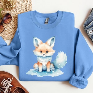 May include: A light blue sweatshirt with a watercolor illustration of a cute orange fox with a white tail. The fox is sitting on a blue background with white snowflakes.