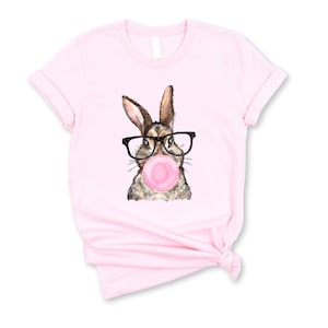 May include: Pink t-shirt with a graphic of a brown rabbit wearing glasses and blowing a pink bubble.