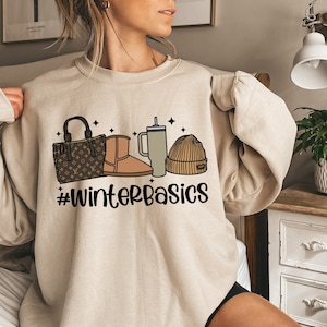 May include: A beige sweatshirt with a graphic design of a handbag, boots, a coffee cup, and a beanie. The graphic is accompanied by the text "#WinterBasics".