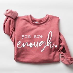 May include: A pink sweatshirt with the text "you are enough" in white lettering. The sweatshirt has a small heart symbol next to the text.