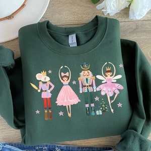 May include: A dark green sweatshirt with a festive design featuring a nutcracker, a ballerina, and a mouse king. The characters are illustrated in a whimsical style with pink, gold, and white accents.