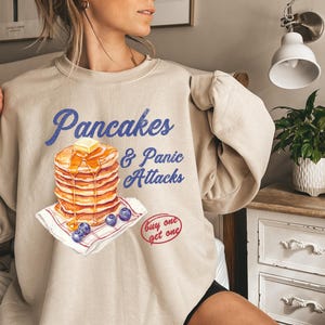 May include: Beige sweatshirt with a graphic of a stack of pancakes with blueberries and the text "Pancakes & Panic Attacks" and "Buy one get one" in red.