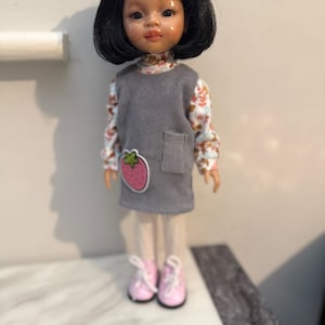May include: A doll with black hair and a cream-colored bow. The doll wears a gray jumper dress with a strawberry patch, a floral print shirt, cream tights, and pink sneakers.