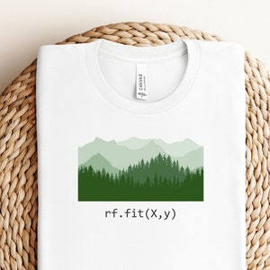 May include: White t-shirt with a green mountain and forest graphic. The design features layers of green trees and mountains, with the text "rf.fit(X,y)" below the graphic. The shirt is folded on a woven surface.
