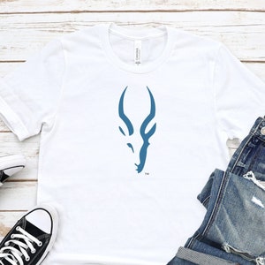 May include: A white t-shirt with a blue stylized antelope head graphic. The shirt is laid flat on a white wood background with a pair of black canvas shoes and blue jeans.