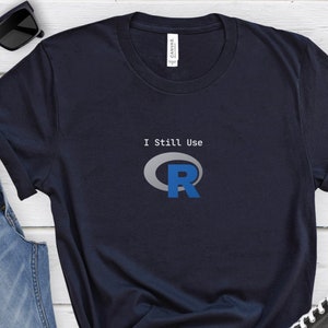 May include: Navy blue t-shirt with the text "I Still Use R" and a graphic of the letter "R" in blue. The shirt is styled with blue jeans, black sunglasses, and black Converse sneakers.
